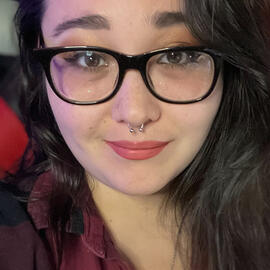 Selfie of a femme with dark brown hair and black thick-rimmed glasses smiling slightly.
